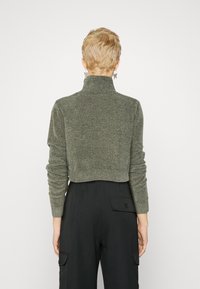 Przycięty, ciemnozielony sweter z dzianiny z wysokim kołnierzem, długimi rękawami i teksturowanym wykończeniem. Noszony z czarnymi spodniami z wysokim stanem, posiadającymi kieszenie.