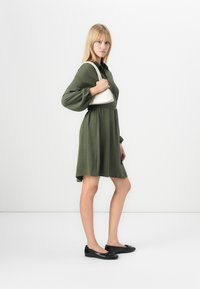 Robe vert olive longueur genoux avec de longues manches bouffantes et une taille cintrée, associée à des ballerines noires et un sac à main blanc. Texture lisse.