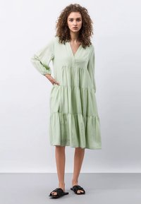 Vaaleanvihreä, kerrostettu mekko V-pääntiellä ja pitkillä hihoilla, valmistettu pehmeästä kankaasta. Ominaisuudet rento istuvuus ja hienovarainen tekstuuri.