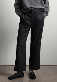 Wijduitlopende broeken van donkergrijze stof met een subtiele textuur, met een hoge taille en omgevouwen zoom. Gepaard met zwarte loafers en grijze sokken.