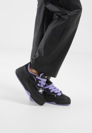 Sorte og lilla sneakers med vedhæng af tegneseriefigur båret med korte sorte bukser, fod lettet lidt fra hvidt gulv.
