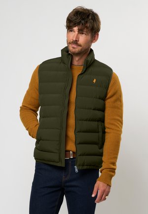 Gilet matelassé kaki avec un col haut, comportant des sections cousues, une fermeture éclair et un petit logo brodé orange. Porté par-dessus un pull moutarde.