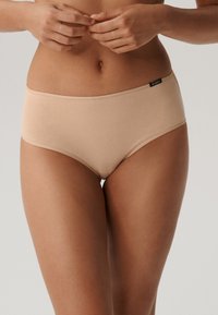 Beżowe bawełniane majtki typu boyshort o gładkiej fakturze, średnio wysokim kroju i małej czarnej metce przy pasku dla oznaczenia marki.