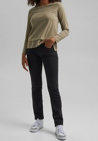Olivenfarbenslangärmliges Oberteil mit geschichtetem Saum, kombiniert mit schwarzen Skinny-Jeans und weißen High-Top-Sneakern. Glattes Material, lässige Passform, keine Muster.