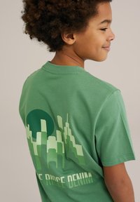 Žalias medvilnės marškinėlis su miesto silueto grafika ir apvaliu saule įvairių žalių atspalvių ant nugaros. Apvalus kaklas ir trumpomis rankovėmis.