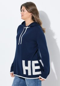 Sweat à capuche marine avec accents blancs et texture tricotée, comportant un grand "HEY" à l'ourlet, des manches longues et une coupe droite.