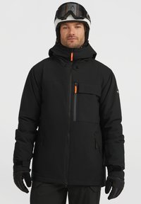 Zwarte skijack met een capuchon, voorzien van oranje accenten, meerdere zakken en een textuur aan de buitenkant. Draagt met handschoenen en een helm.