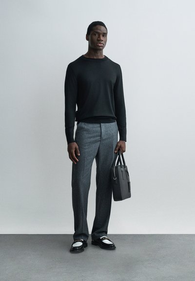 Un homme se tient contre un mur pâle, portant un pull noir, un pantalon gris sur mesure, des mocassins noir et blanc, tenant une mallette noire dans la main droite.