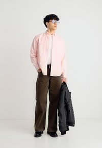 Jeune homme portant des lunettes, chemise rose clair, pantalon marron, tenant une veste noire, debout sur un fond blanc uni.