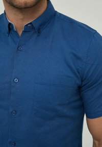 Chemise bleu à manches courtes en tissu lisse, avec un col boutonné et une poche poitrine unique, ornée de boutons assortis.