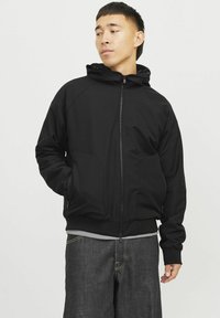 Schwarze Zip-Jacke mit Kapuze, aus glattem Stoff, mit Fronttaschen und geripptem Saum. Getragen über einem hellgrauen Shirt und dunklen Jeans.