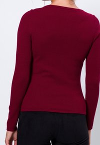 Haut bordeaux à manches longues avec un tissu texturé, un col rond et une coupe ajustée, associé à un pantalon noir, vu de côté.