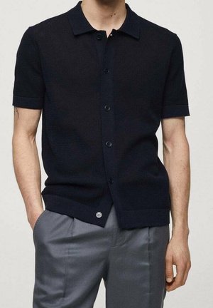 Polo en tricot navy à manches courtes avec un devant à boutons, col, et tissu texturé. Porté avec un pantalon gris.