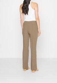 Pantaloni attillati marroni con una consistenza liscia, caratterizzati da una cintura ampia e gambe dritte. Indossati con una maglietta bianca senza maniche e scarpe chiare.