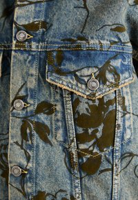 Denim jakna z modro podlago in temnimi cvetličnimi vzorci, srebrnimi gumbi ter kontrastnim šivanjem na prsnem žepu in šivih.