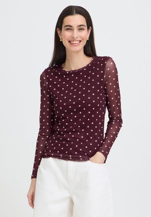 Uśmiechnięta kobieta w długim, prześwitującym bordowym topie w różowe kropki i białych spodniach, z ręką w kieszeni, na jednolitym tle.