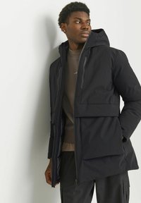 Sort hættejakke lavet af vandafvisende materiale med frontlynlås, store lommer og en glat tekstur. Casual design og tætsiddende pasform.