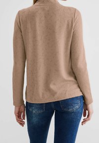 Maglia beige a maniche lunghe con colletto, abbinata a jeans in denim blu. La maglia presenta un motivo sottile e una vestibilità rilassata.