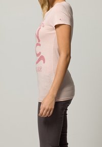 Světle růžové bavlněné tričko s přiléhavým střihem a krátkými rukávy. Na přední straně má stylizovanou grafiku v tmavší růžové barvě.