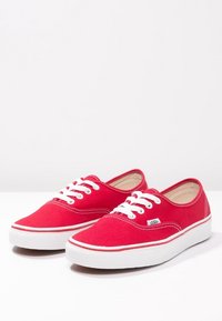 Rote Canvas-Sneaker mit weißen Gummisohlen, ausgestattet mit weißen Schnürsenkeln und einem kleinen Logokennzeichen an der Seite. Klassisches Low-Top-Design.