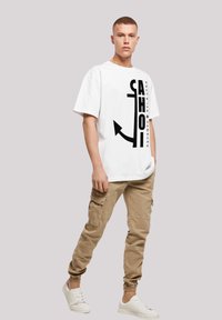 White oversized t-shirt with black anchor graphic and text, paired with beige cargo pants featuring side pockets, and white sneakers.