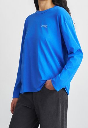 Personne portant une chemise bleu vif à manches longues avec le texte « OBEY » sur la poitrine, pantalon noir, main dans la poche, fond blanc.