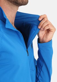 Giacca a pullover con zip di colore blu, foderata con un tessuto dalla texture, dotata di colletto e hardware nero. Il materiale appare liscio con motivi geometrici.