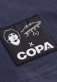 Zwarte stoffen label met een witte silhouet van een persoon, handtekening en de tekst "x COPA" op een donkerblauwe kledingachtergrond.