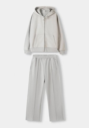 Hellgrauer Sweatshirt mit einem Reißverschluss vorne, Kapuze mit Kordelzug und zwei Taschen, kombiniert mit passenden hellgrauen Hosen mit elastischem Bund und seitlichen Taschen.