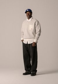 Grauer Sweatshirt mit einer Vordertasche, über einem weißen Hemd getragen. Schwarze lässige Hose und schwarze Schuhe. Blauer Baseballcap.