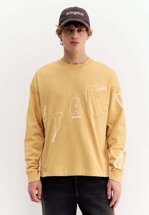 Jeune homme portant une chemise à manches longues beige avec un texte blanc et une poche poitrine, un jean noir et une casquette foncée avec des inscriptions blanches.