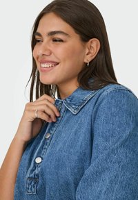 Denimová košile ve středně modré barvě s límcem na knoflíky, vybavená kovovými knoflíky a texturovanou tkaninou. Model jemně drží límec.