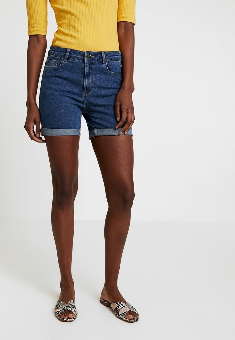 Personne portant un haut jaune, un short en jean bleu retroussé et des sandales ouvertes à motif serpent, debout devant un fond uni.