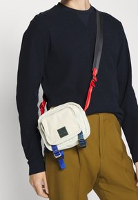 Personne portant une chemise à manches longues bleu marine foncé, un pantalon moutarde et un petit sac blanc porté en bandoulière avec des sangles ajustables rouges et bleues.