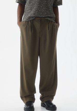 Person trägt lockere olivgrüne Hose, ein grau strukturiertes kurzärmeliges Oberteil und schwarze Schuhe mit klobiger Sohle, Hände in den Taschen.