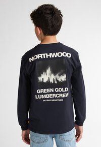 Maglietta a maniche lunghe colore blu navy con stampa grafica bianca contenente il testo "NORTHWOOD" e "GREEN GOLD LUMBERCREW", insieme a una foto di una foresta. Materiale in cotone.