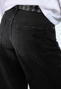 Čierne džínsy z denimu s zadným vreckom a ošúchanými detailmi, doplnené čiernym koženým opaskom so striebornými očkami.