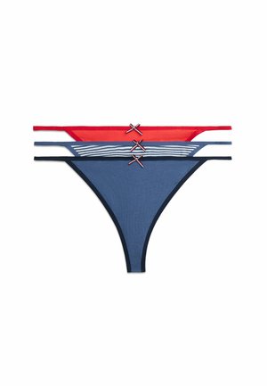 Ung kvinde står barfodet med armene over kors, iført en hvid tanktop og rødt undertøj, smilende mod en ensartet hvid baggrund.
