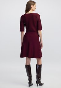 Bordeaux breipak met korte mouwen en een uitlopende rok, gecombineerd met hoogglanzende bruine laarzen tot de knie. Minimale naden en een getailleerde silhoët.