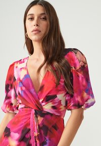 Bloemmotief jurk in levendige roze, rode en blauwe tinten. Heeft korte, pofmouwen, een diepe V-hals en een geknoopte taille-detail.