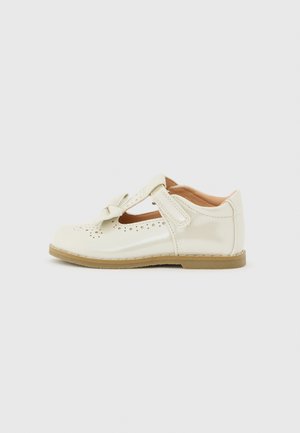 Chaussure pour enfant de type Mary Jane en cuir verni couleur crème avec des détails perforés, un nœud décoratif, une bride en velcro et une semelle plate couleur sable.