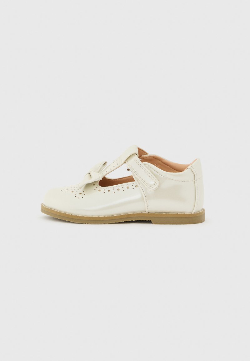 Chaussure pour enfant de type Mary Jane en cuir verni couleur crème avec des détails perforés, un nœud décoratif, une bride en velcro et une semelle plate couleur sable.