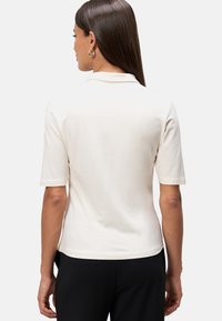 Crèmegekleurde poloshirt met korte mouwen en een zachte textuur. Voorzien van een klassieke kraag en een rechte zoom, gecombineerd met zwarte broek.