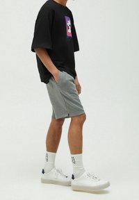 T-shirt noir à manches courtes avec design graphique, associé à un short gris. Des baskets blanches avec des accents bleu marine et des chaussettes côtelées complètent la tenue.