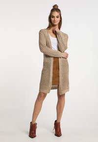 Cardigan beige lavorato a maglia con un design a trama aperta, maniche lunghe e lunghezza sotto il ginocchio. Abbinato a una gonna in suede marrone e stivali alla caviglia.