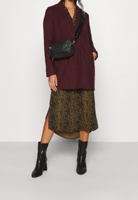 Bordeauxwollen jas met een reverskraag en zijzakken, over een zwart-gouden jurk met patroon. Zwarte lederen crossbodytas en laarzen tot de knie.