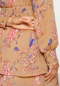 Vestido floral em tecido bege claro, com padrões de flores em vermelho e azul, cintura franzida, e mangas longas com detalhes nos punhos.