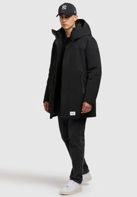 Joven vestido con una chaqueta de invierno con capucha negra, jeans negros, zapatillas blancas y una gorra de béisbol negra, de pie contra un fondo gris claro.
