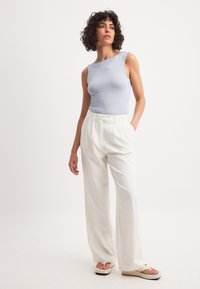 Svetlo modra brezrokavna majica v kombinaciji z belimi hlačami visoko pasu; gladka tkanina z sproščenim krojem. Ravne sandale z debelim podplatom.