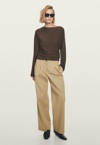Persona che indossa occhiali da sole neri, un top a maniche lunghe marrone scuro, pantaloni beige a gamba larga e scarpe nere con punta, in piedi con una mano in tasca.